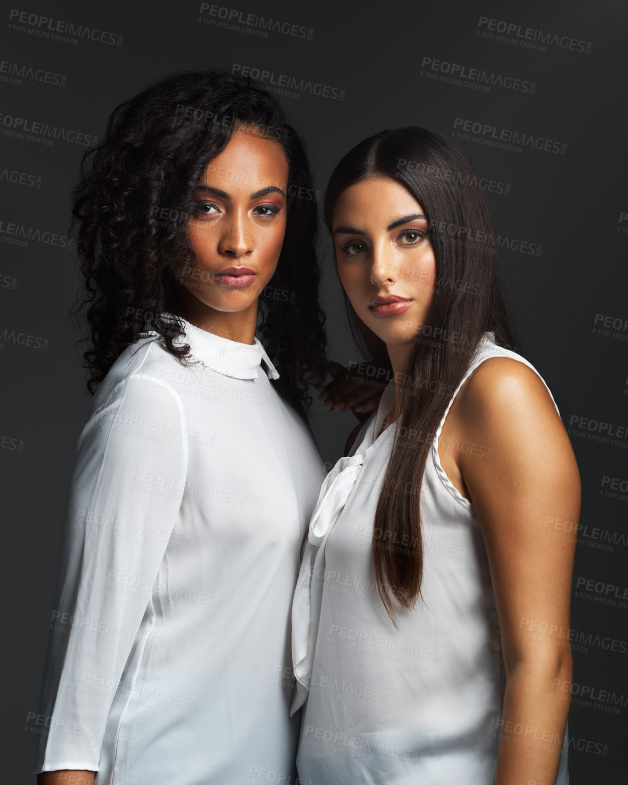 Buy stock photo Portrait of two attractive young women wearing white blouses posing closely together against a dark background in the studio