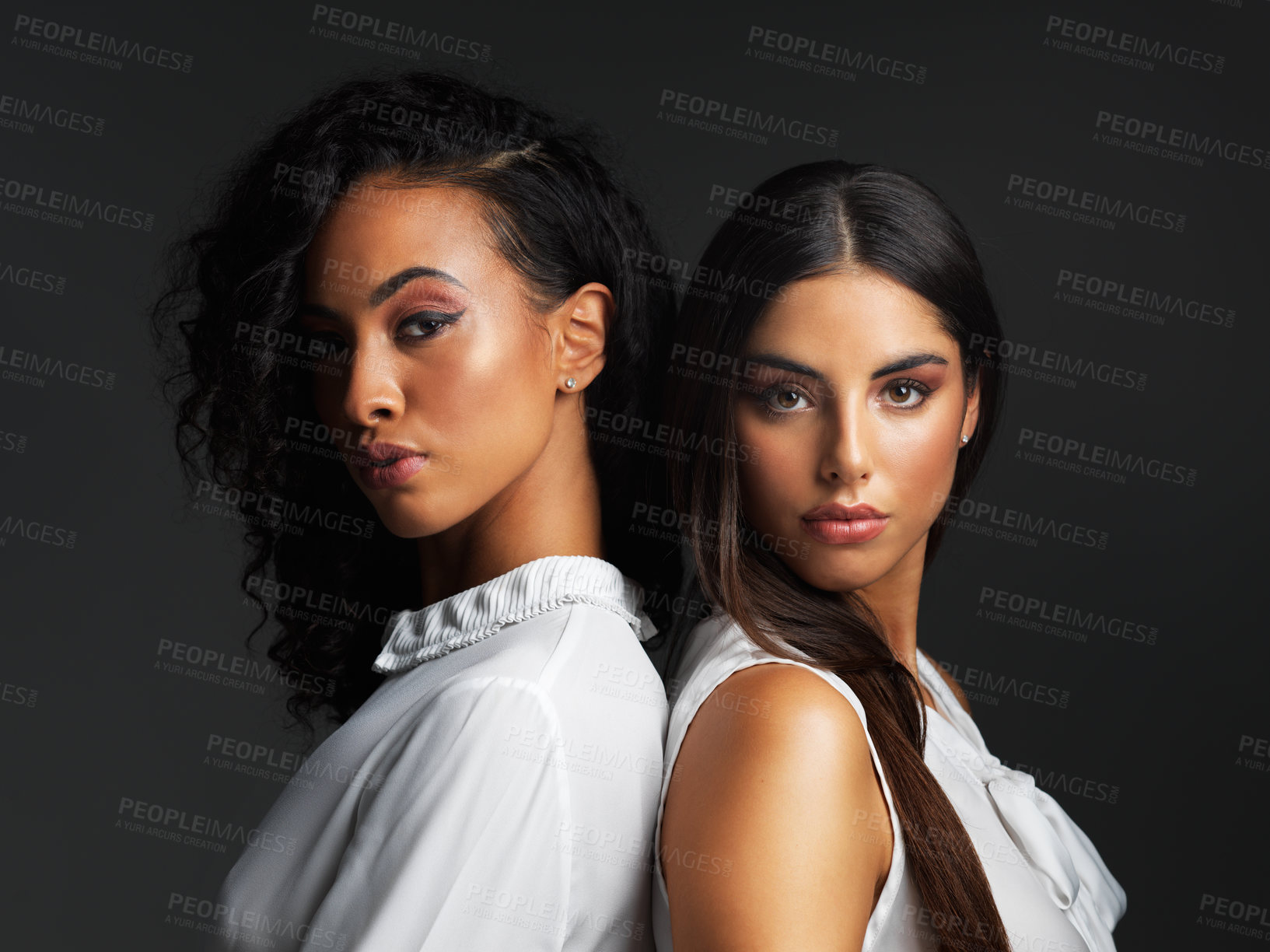 Buy stock photo Portrait of two attractive young women posing back to back against a dark background in a studio