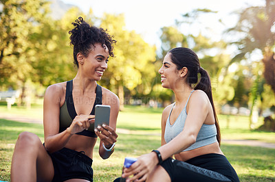 Buy stock photo Cropped shot of two attractive young women sitting next to each other while using a cellphone in the park