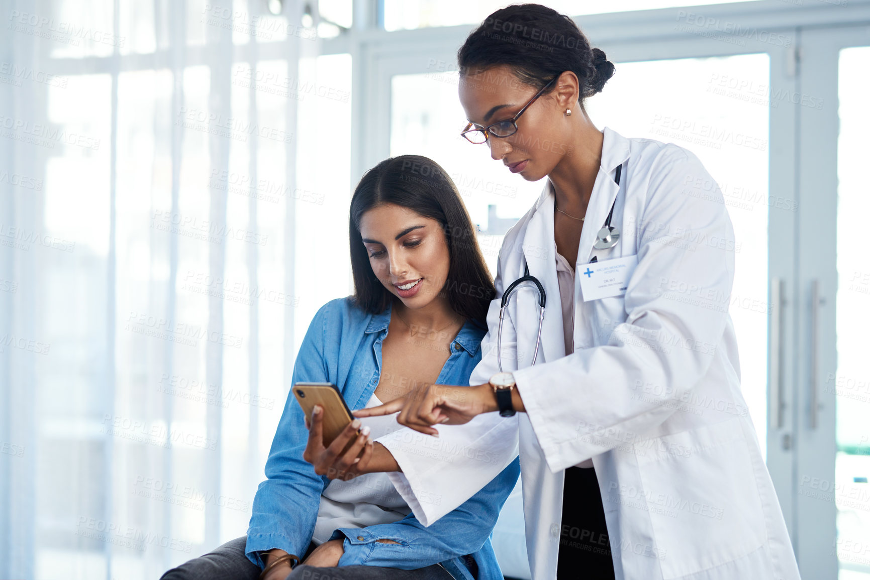Buy stock photo Shot of a young doctor using a smartphone during a consultation with her patient