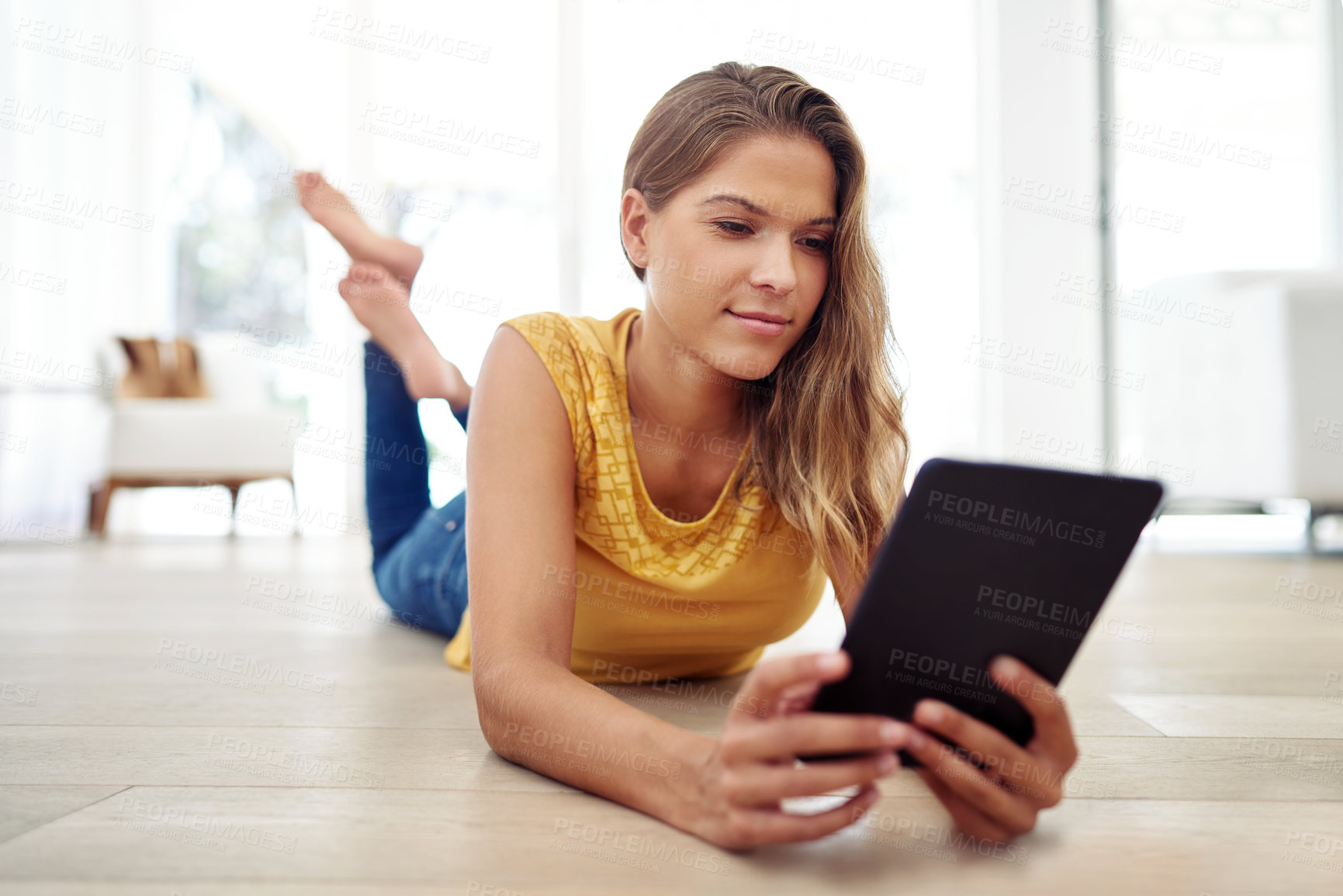 Buy stock photo Full body shot of an attractive young woman sitting and using a tablet in the living room during the day