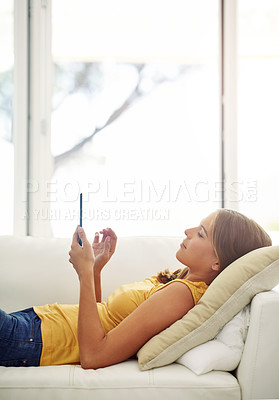 Buy stock photo Cropped shot of an attractive young woman using a digital tablet while lying on a sofa at home