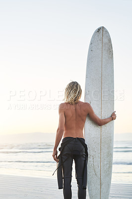 Buy stock photo Rearview shot of a young surfer standing next to his surfboard at the beach