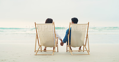 Buy stock photo Rearview shot of an affectionate young couple sitting next to each other and holding hands while on the beach