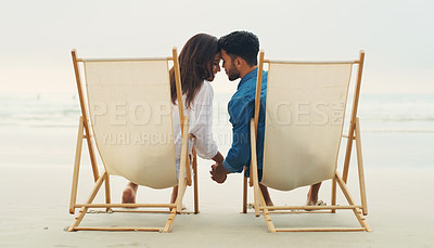 Buy stock photo Rearview shot of an affectionate young couple sitting next to each other and holding hands while on the beach