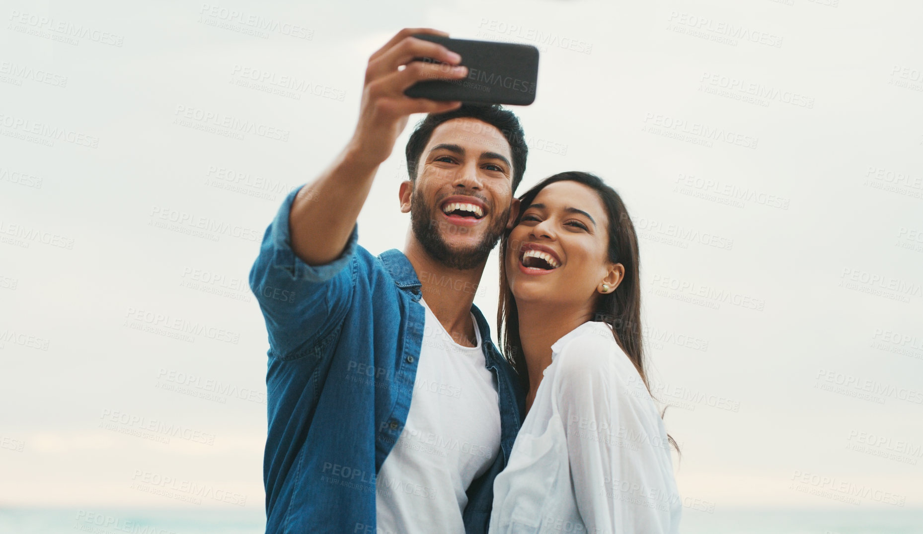 Buy stock photo Cropped shot of a happy young couple posing for a selfie while on the beach during the day