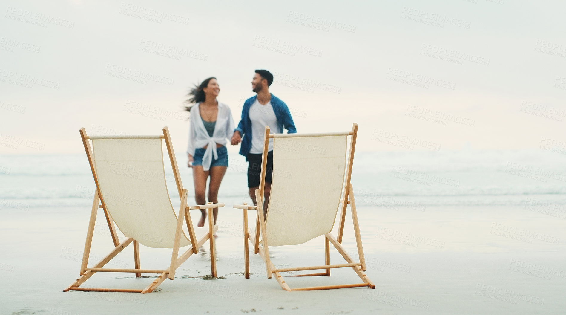 Buy stock photo Full length shot of an affectionate young couple walking towards a set of beach chairs during the day