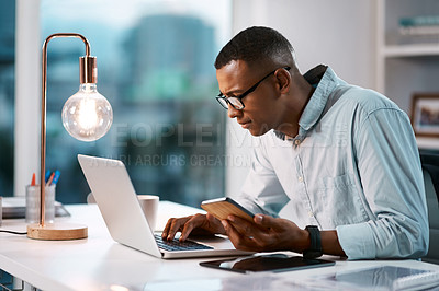 Buy stock photo Shot of a handsome young businessman using his laptop and cellphone while working late in his office