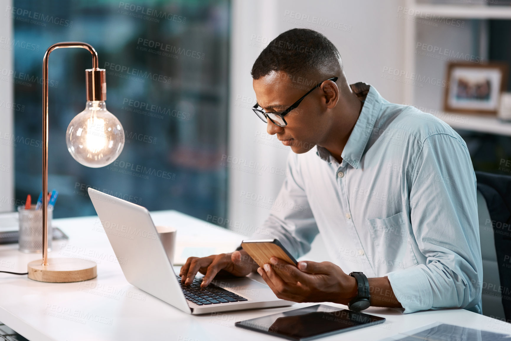 Buy stock photo Shot of a handsome young businessman using his laptop and cellphone while working late in his office