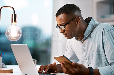 Buy stock photo Shot of a handsome young businessman using his laptop and cellphone while working late in his office