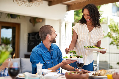 Buy stock photo Shot of a beautiful young woman dishing up salad on her husband's plate while enjoying a meal with family outdoors