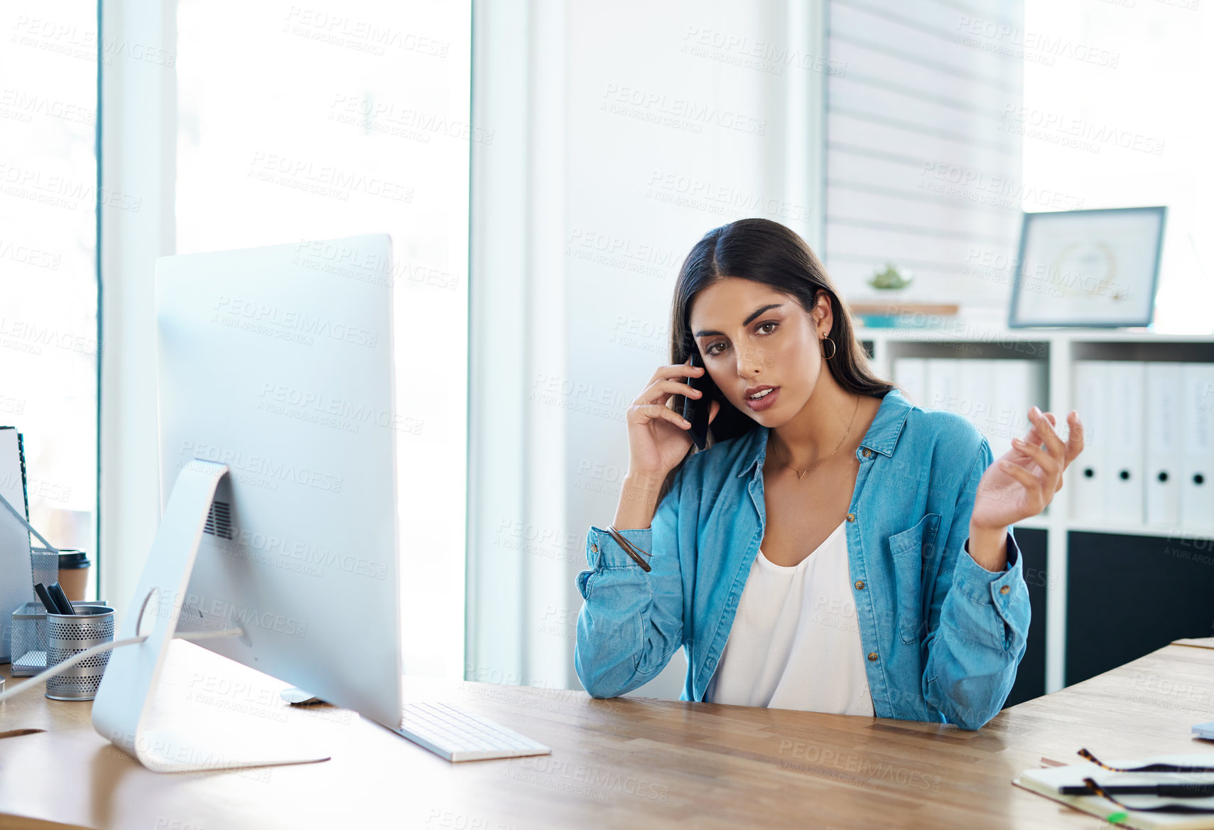 Buy stock photo Shot of a young businesswoman talking on a cellphone while working on a computer in an office