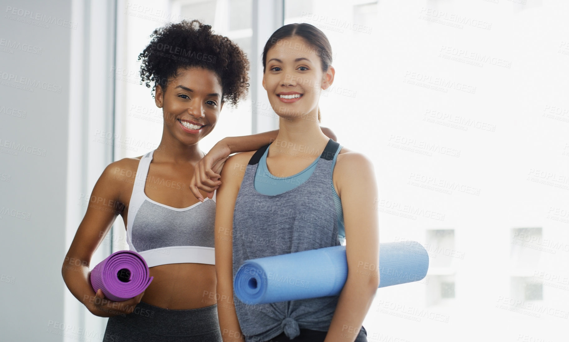 Buy stock photo Cropped portrait of two attractive young women standing and holding yoga mats and posing before working out indoors