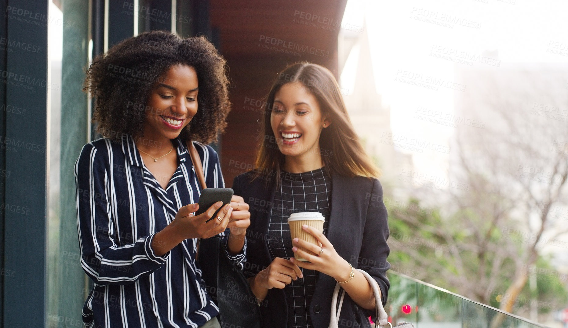 Buy stock photo Cropped shot of two attractive young businesswomen using a cellphone while walking outside during the day