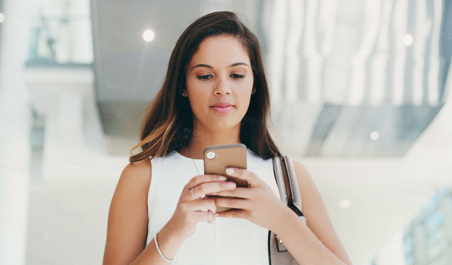 Buy stock photo Shot of a young businesswoman using a smartphone while walking through a modern office