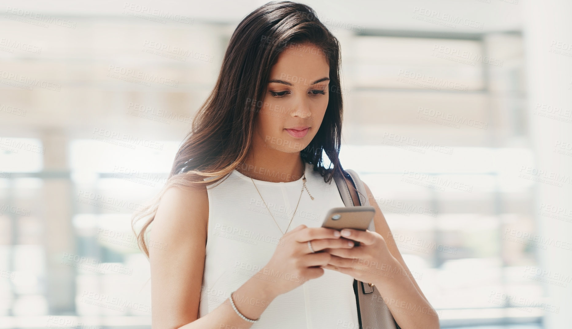 Buy stock photo Shot of a young businesswoman using a smartphone while walking through a modern office