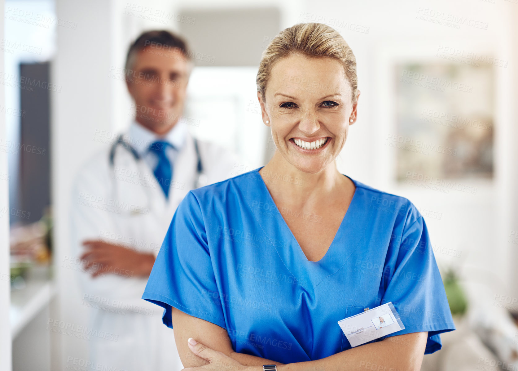 Buy stock photo Portrait of a female nurse standing with her arms crossed in the hospital with a male colleague in the background
