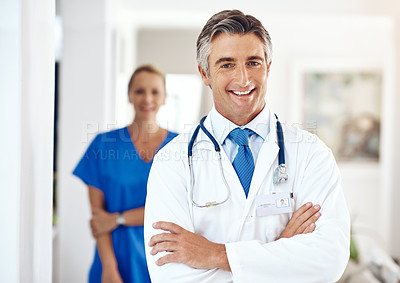 Buy stock photo Portrait of a male doctor standing with his arms crossed in the hospital with a female colleague in a the background