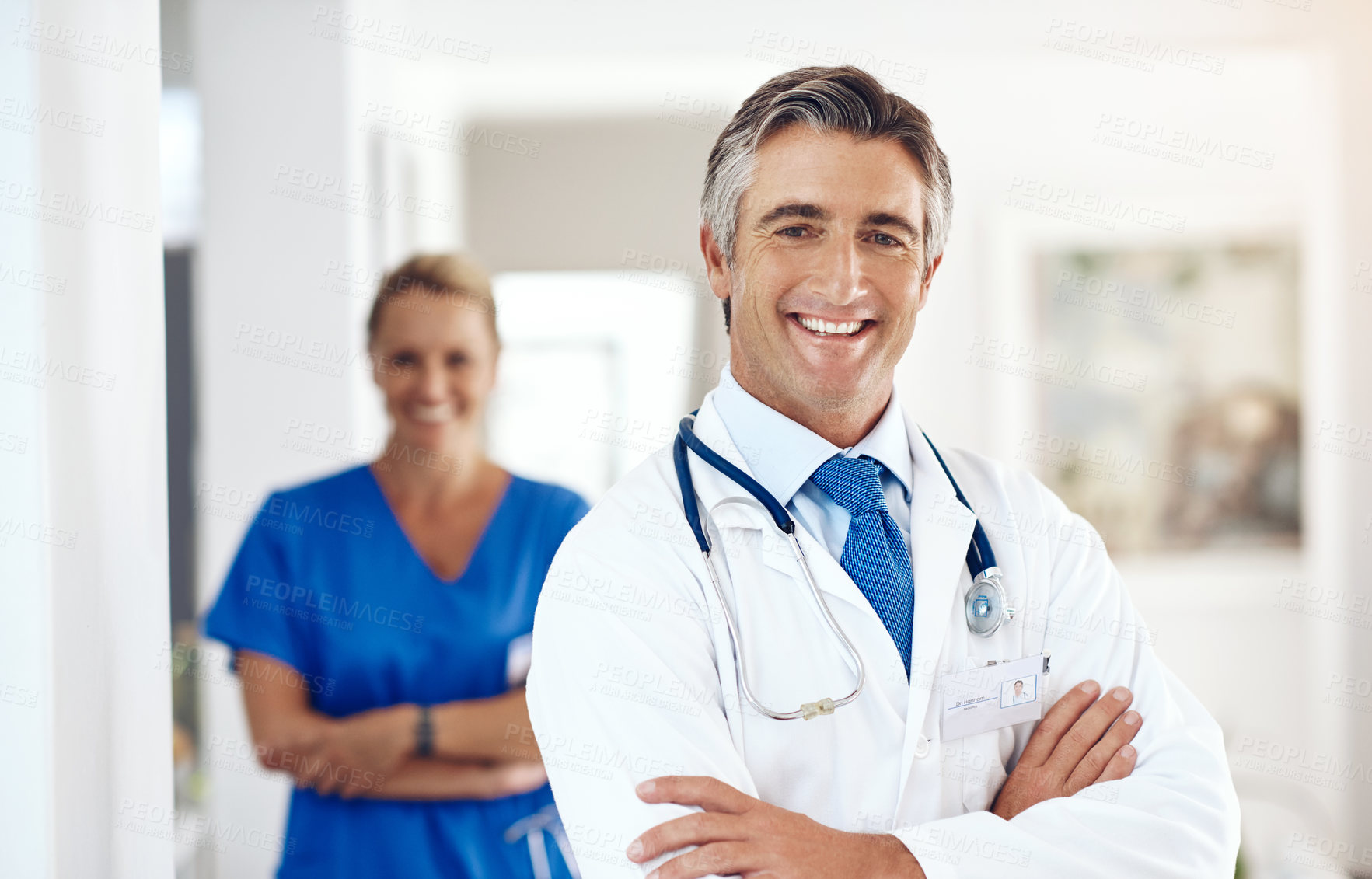 Buy stock photo Portrait of a male doctor standing with his arms crossed in the hospital with a female colleague in a the background