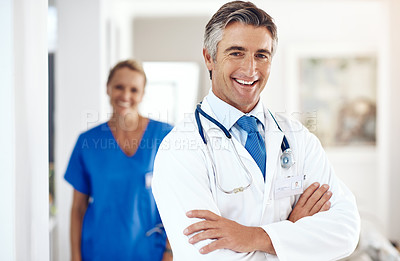 Buy stock photo Portrait of a male doctor standing with his arms crossed in the hospital with a female colleague in a the background