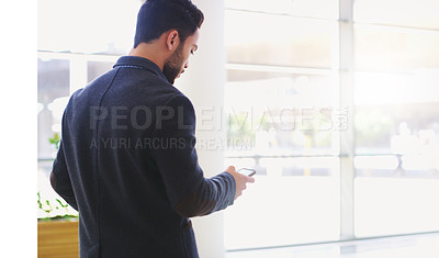 Buy stock photo Rearview shot of a handsome young businessman standing and using his cellphone while pulling a suitcase in the office