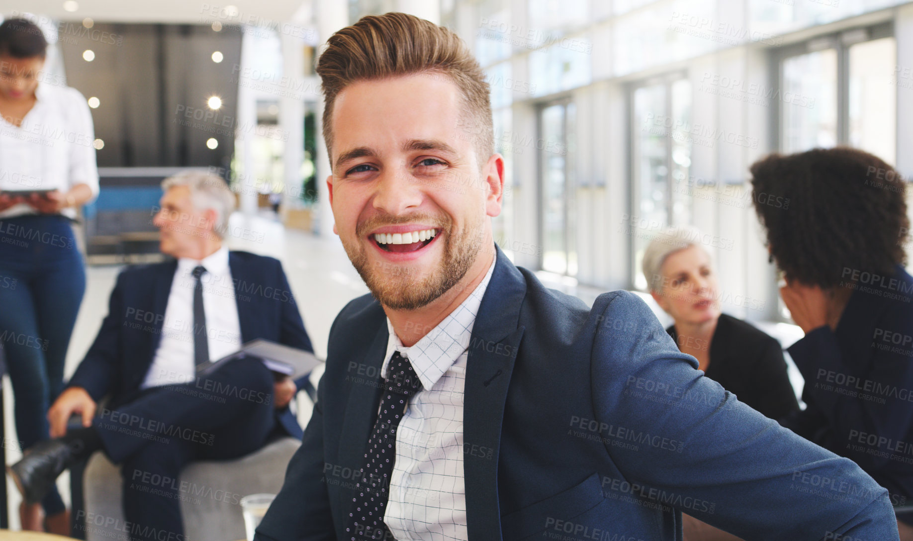 Buy stock photo Cropped portrait of a handsome young businessman sitting and smiling while his colleagues work behind him in the office