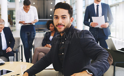 Buy stock photo Cropped portrait of a handsome young businessman sitting and smiling while his colleagues work behind him in the office