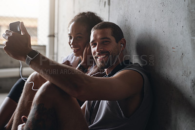Buy stock photo Shot of a sporty young couple sitting down against a wall and using a cellphone while exercising indoors