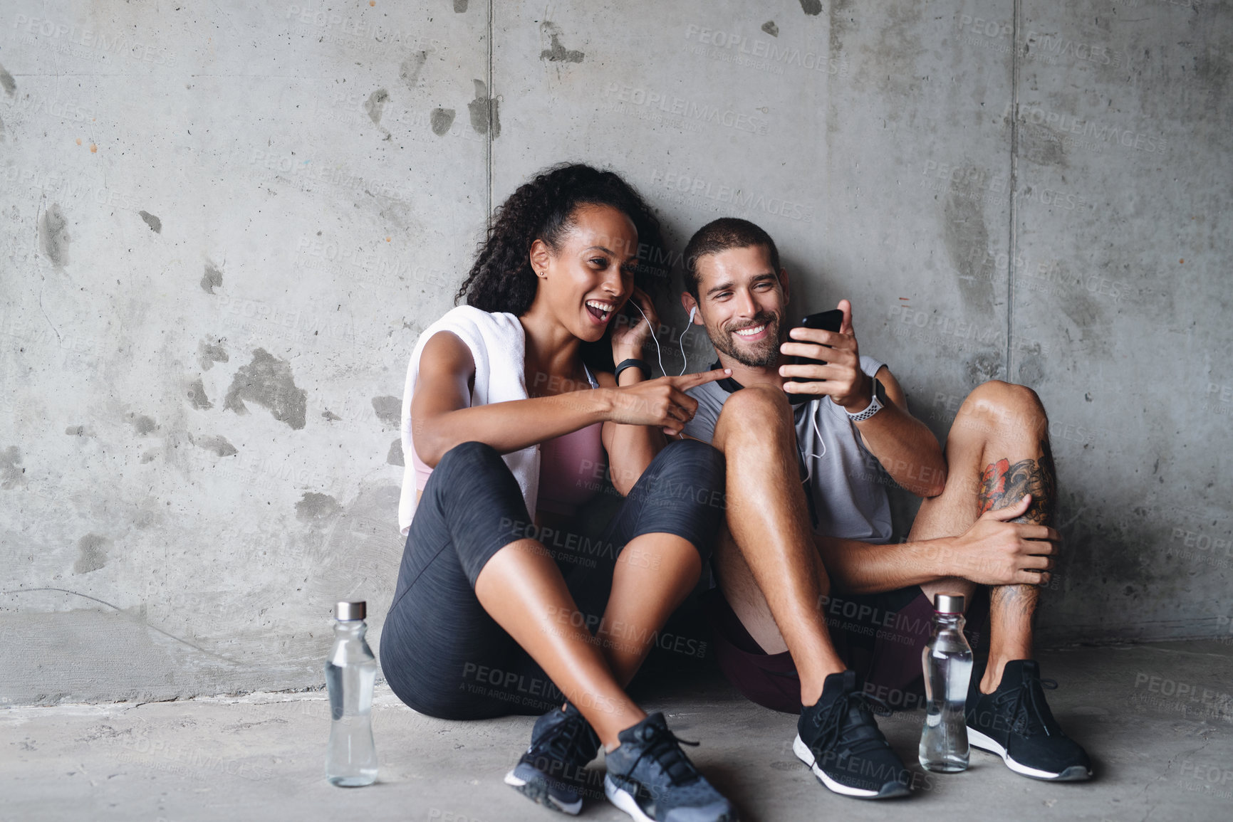 Buy stock photo Shot of a sporty young couple using a cellphone together while  sitting down and resting against a wall
