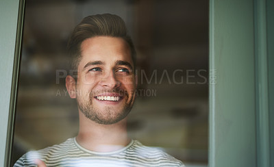 Buy stock photo Shot of a handsome young man looking out of a window at home and smiling