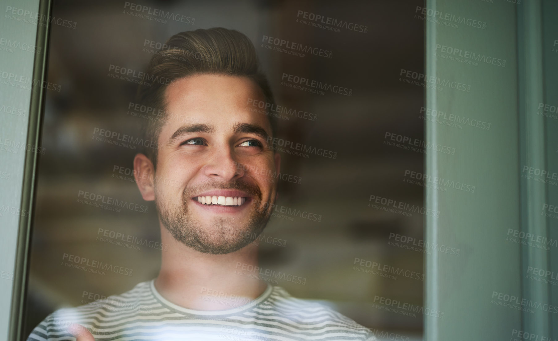 Buy stock photo Shot of a handsome young man looking out of a window at home and smiling