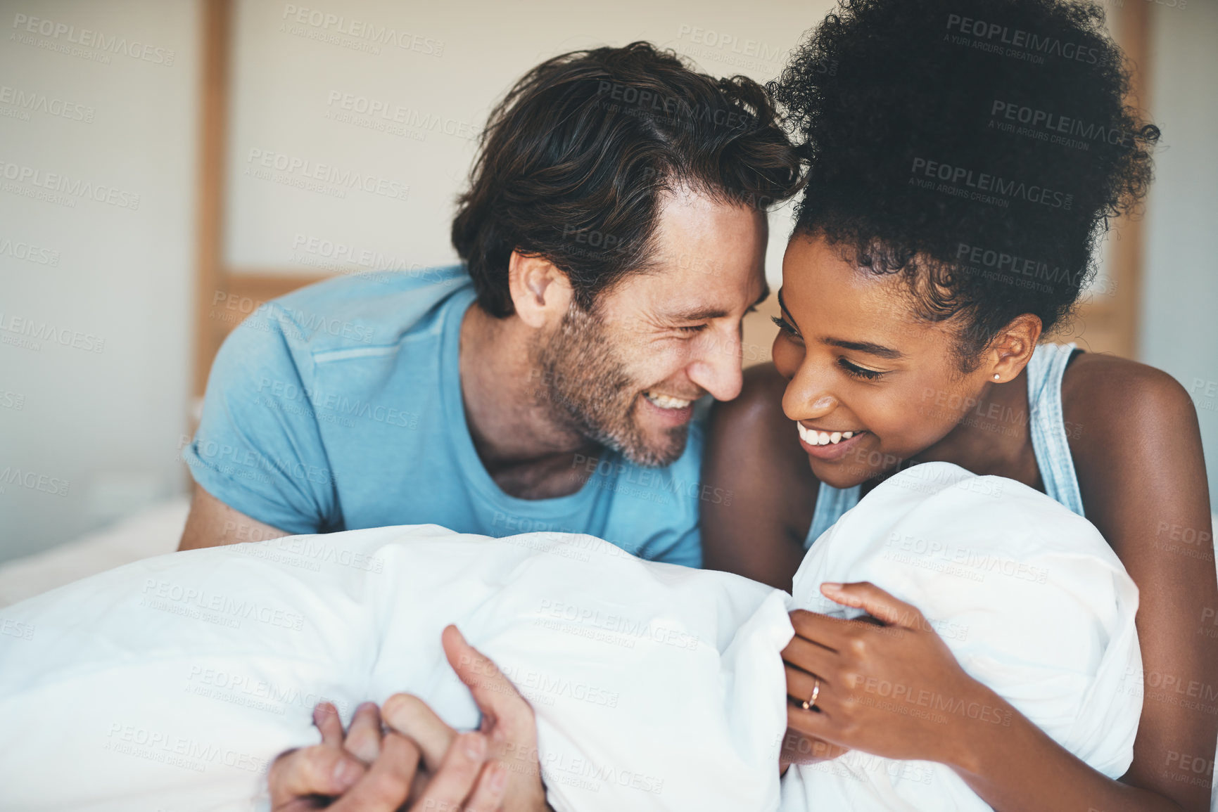 Buy stock photo Happy, loving and funny interracial couple smiling and laughing while lying in bed and sharing a tender moment. Carefree husband and wife relaxing and having fun while bonding in the bedroom