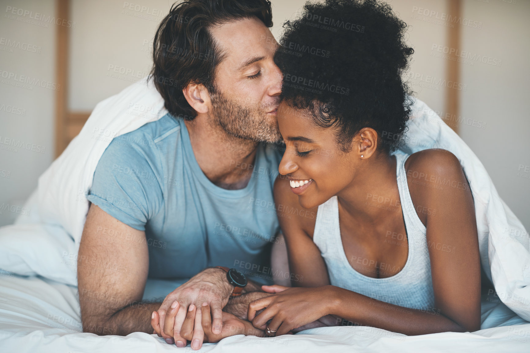 Buy stock photo Happy, carefree and loving couple share a kiss lying in bed together in the morning. Interracial, intimate and caring husband and wife bonding and showing affection while bonding together at home