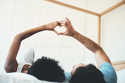 Buy stock photo Interracial couple, hands and heart emoji in bed for morning, love or care relaxing together at home. Man and woman lying in bedroom with touching hand for loving symbol, sign or gesture at the house