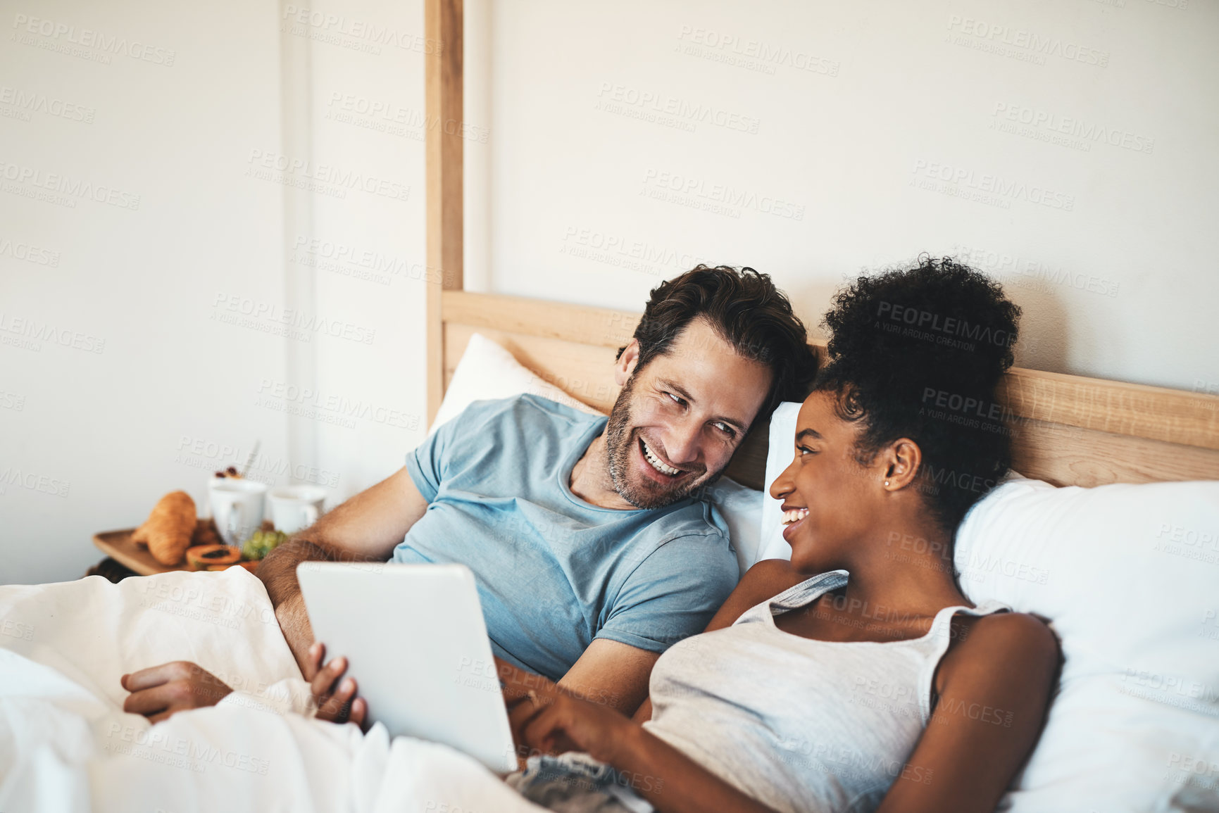 Buy stock photo Happy couple, tablet and laughing on bed for joke, funny entertainment or online streaming at home. Interracial man and woman person on technology in relax for morning or social media in the bedroom