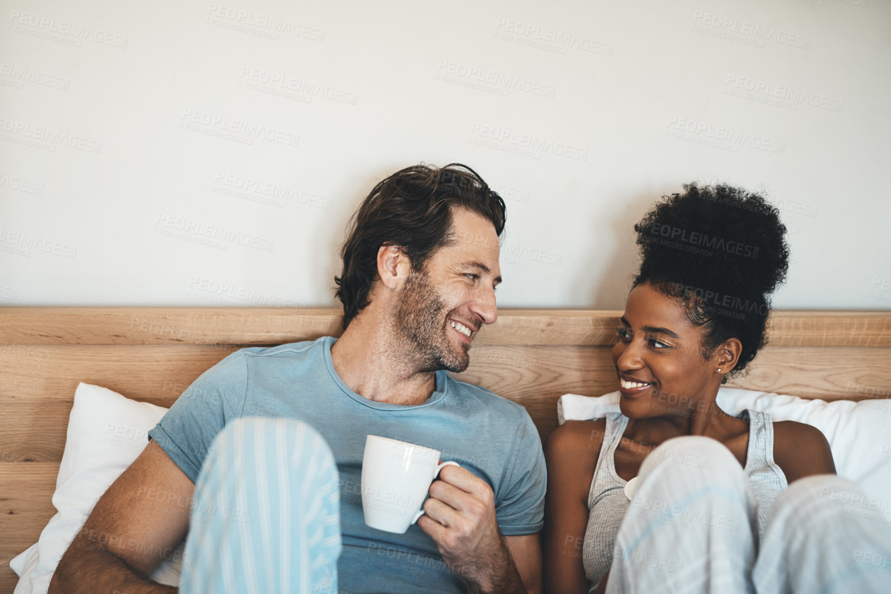 Buy stock photo Happy young interracial couple having a cup of coffee on a bed in the morning at home. Romantic people in love, smiling in the bedroom with a warm beverage with loving man and woman in happiness.