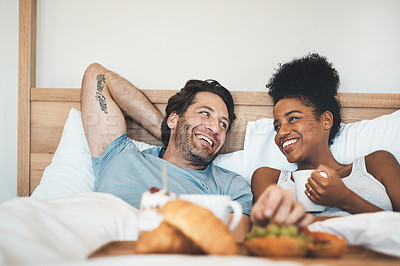 Buy stock photo Happy interracial couple, bed and breakfast in relax for morning, bonding or relationship at home. Man and woman smiling with food, coffee or meal relaxing on holiday or weekend together in bedroom