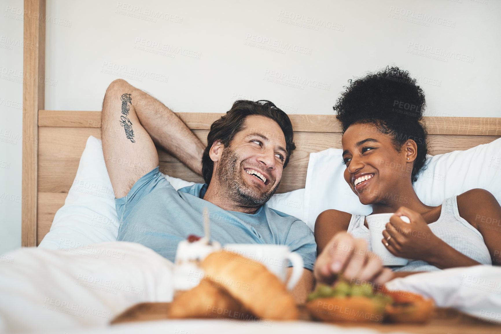 Buy stock photo Happy interracial couple, bed and breakfast in relax for morning, bonding or relationship at home. Man and woman smiling with food, coffee or meal relaxing on holiday or weekend together in bedroom