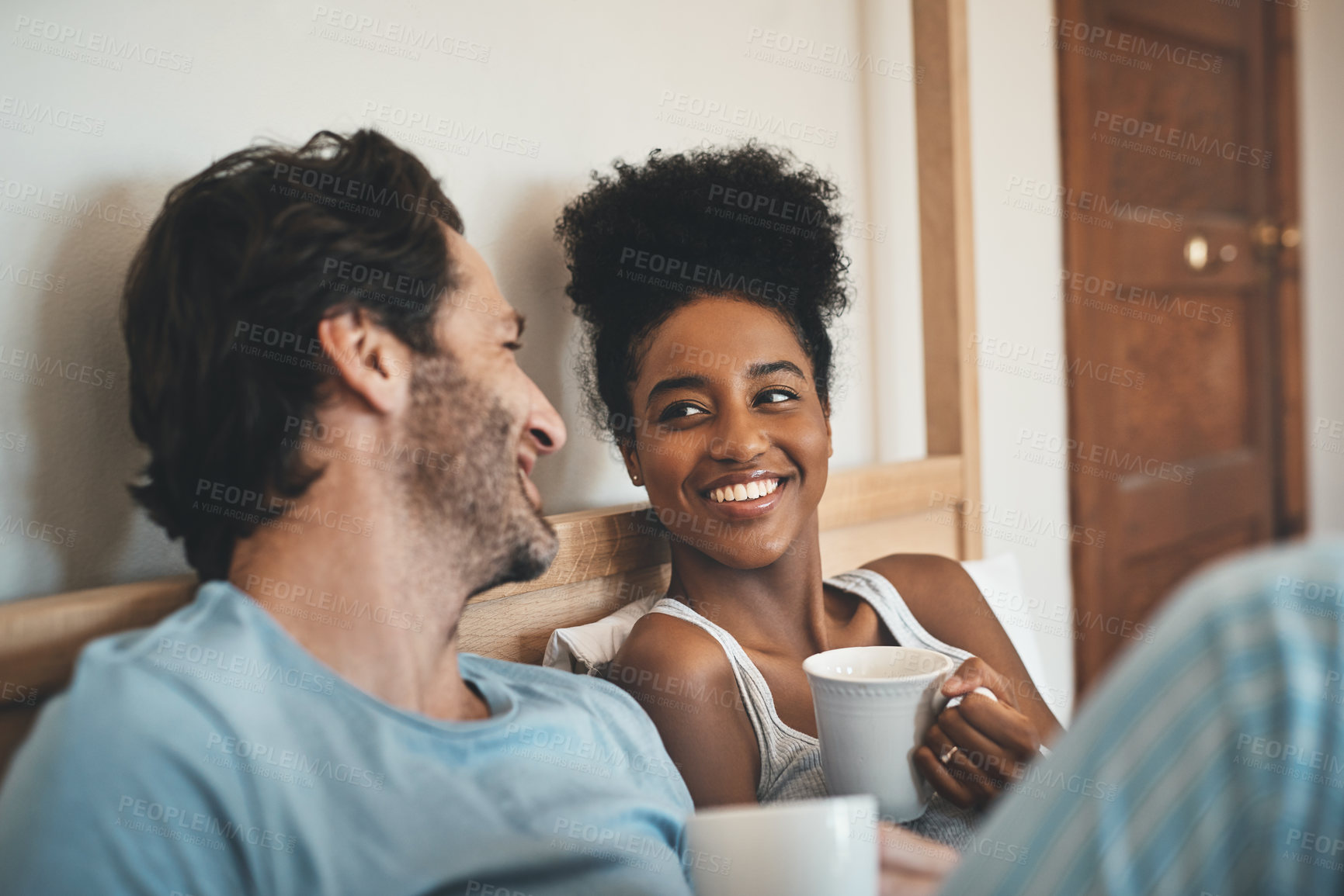 Buy stock photo Romantic, loving and happy interracial couple having a cup of coffee on a bed in the morning at home. People in love, smiling in the bedroom with a warm beverage. Happy young man and woman bonding