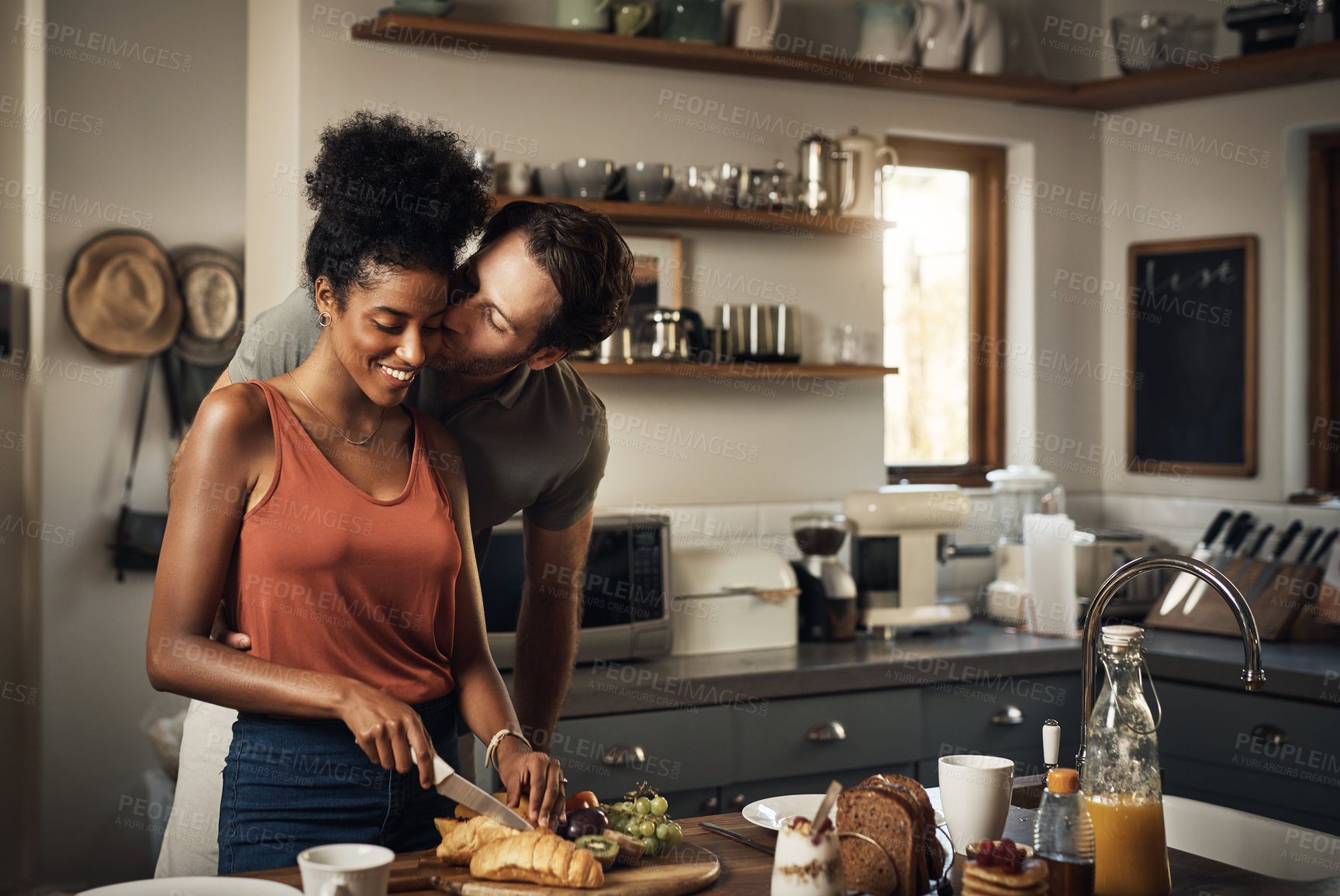 Buy stock photo Interracial couple, kiss and cooking in kitchen for morning breakfast, love or caring relationship at home. Man kissing woman while making food, romantic meal or cutting ingredients on table in house