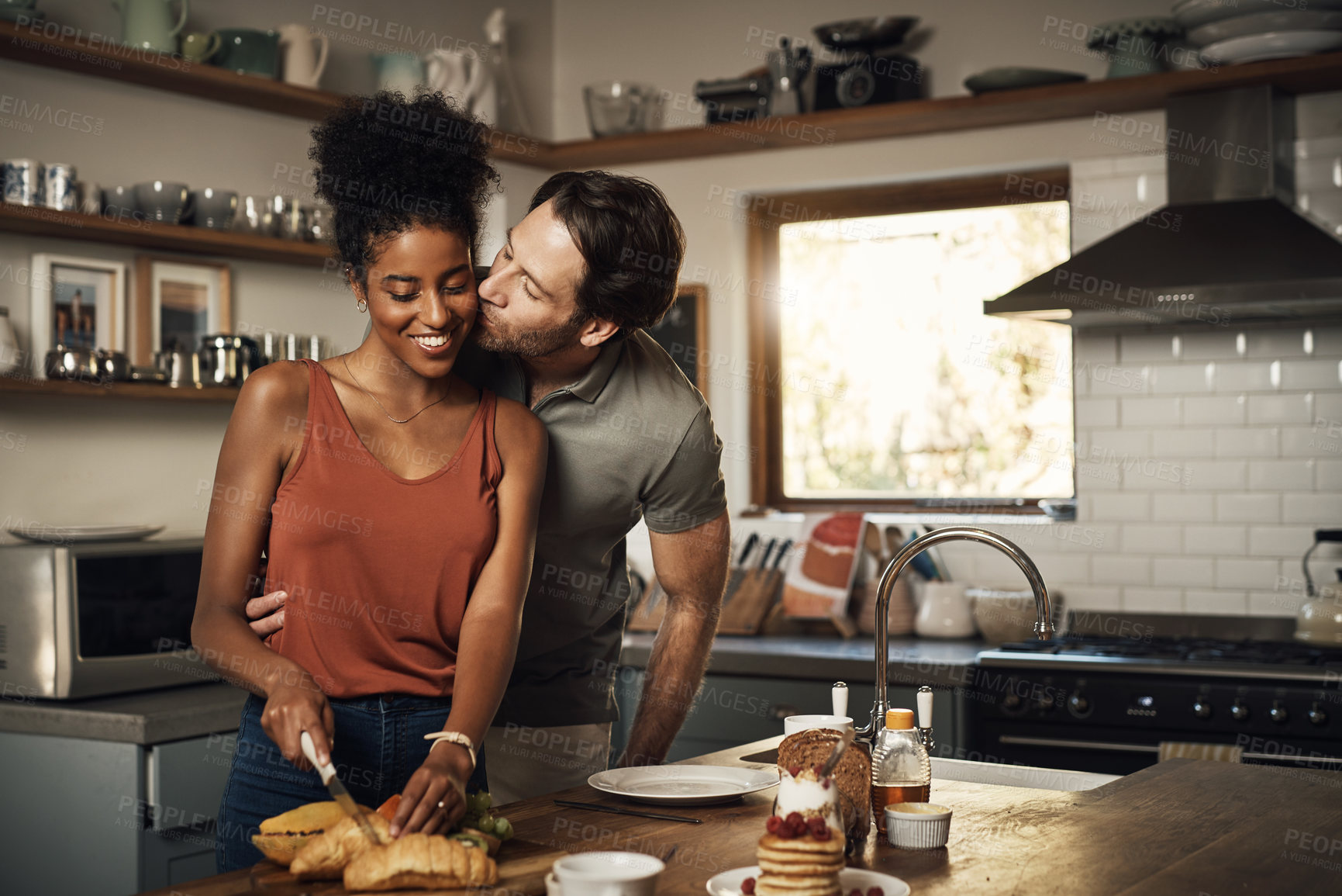 Buy stock photo Interracial couple, kiss and hug in kitchen for cooking, morning breakfast, love or caring relationship at home. Man kissing woman preparing food, meal or cutting ingredients on table at the house