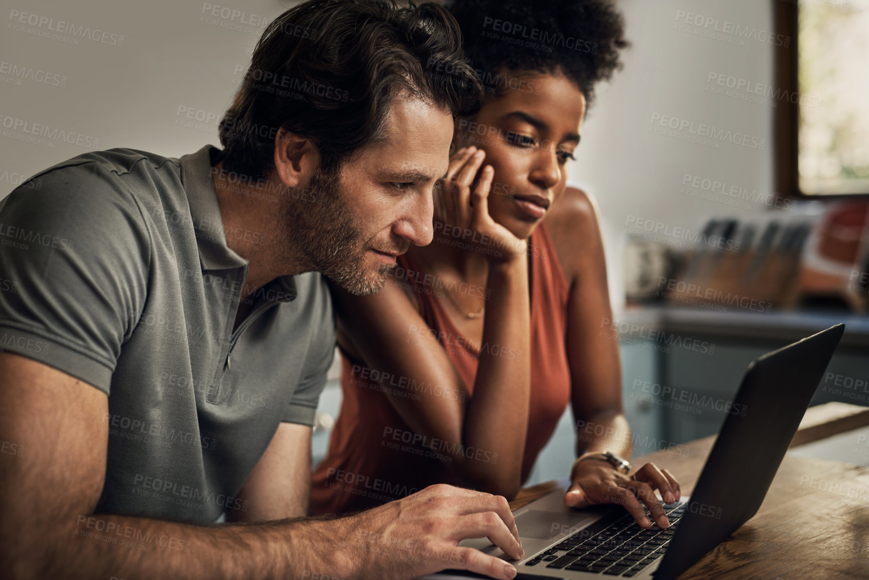 Buy stock photo Diverse couple searching for a new rental apartment home on their laptop sitting together in their kitchen at their house. Man and woman in an interracial marriage relationship doing online shopping