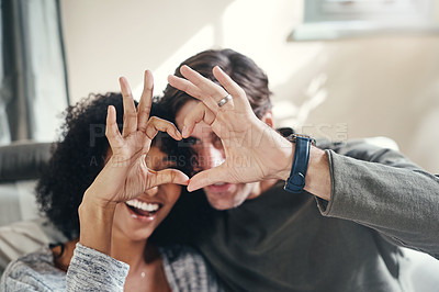 Buy stock photo Shot of a playful young couple making heart shapes with their hands in their living room at home