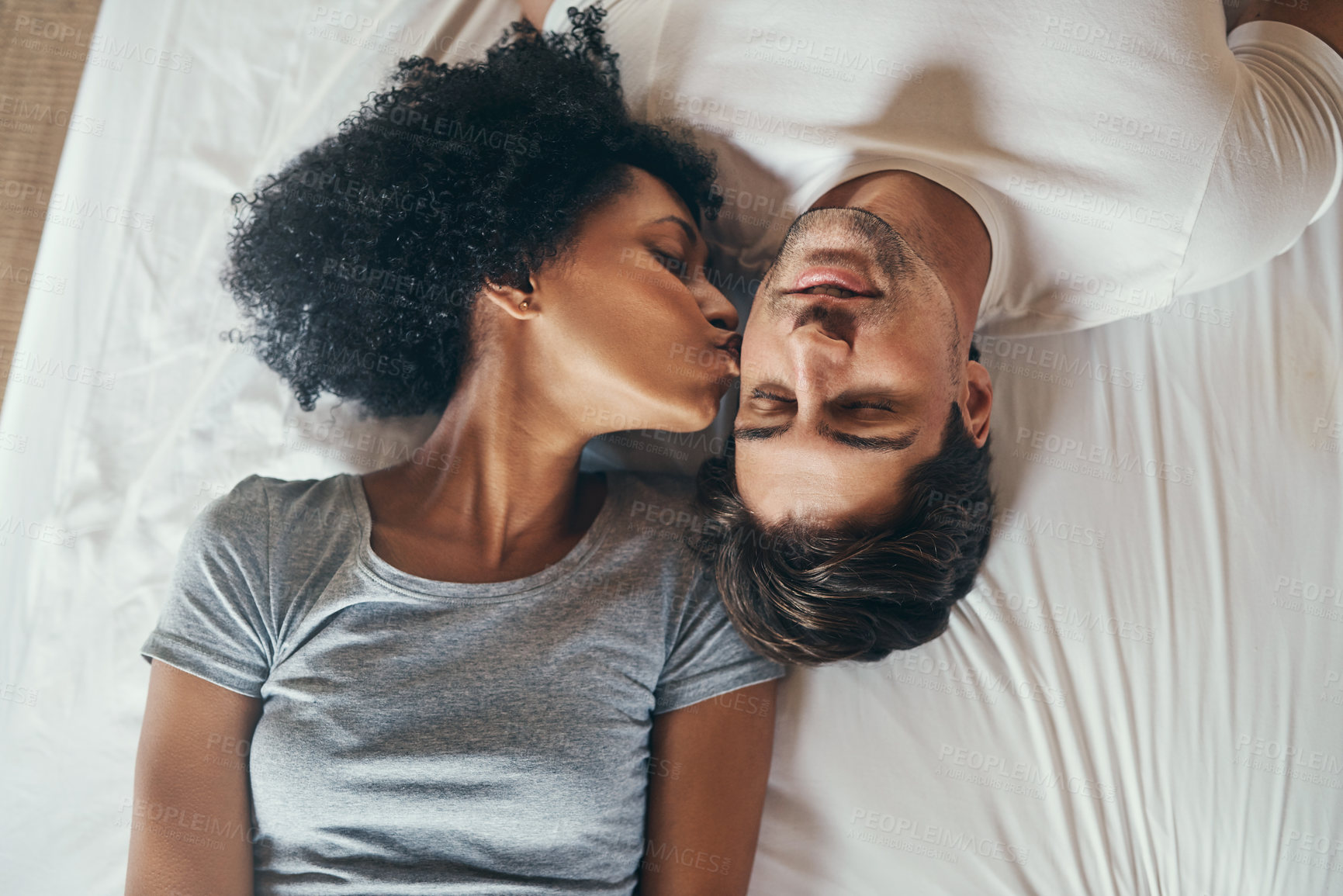 Buy stock photo High angle shot of a young woman kissing her boyfriend on the cheek while lying in bed together at home