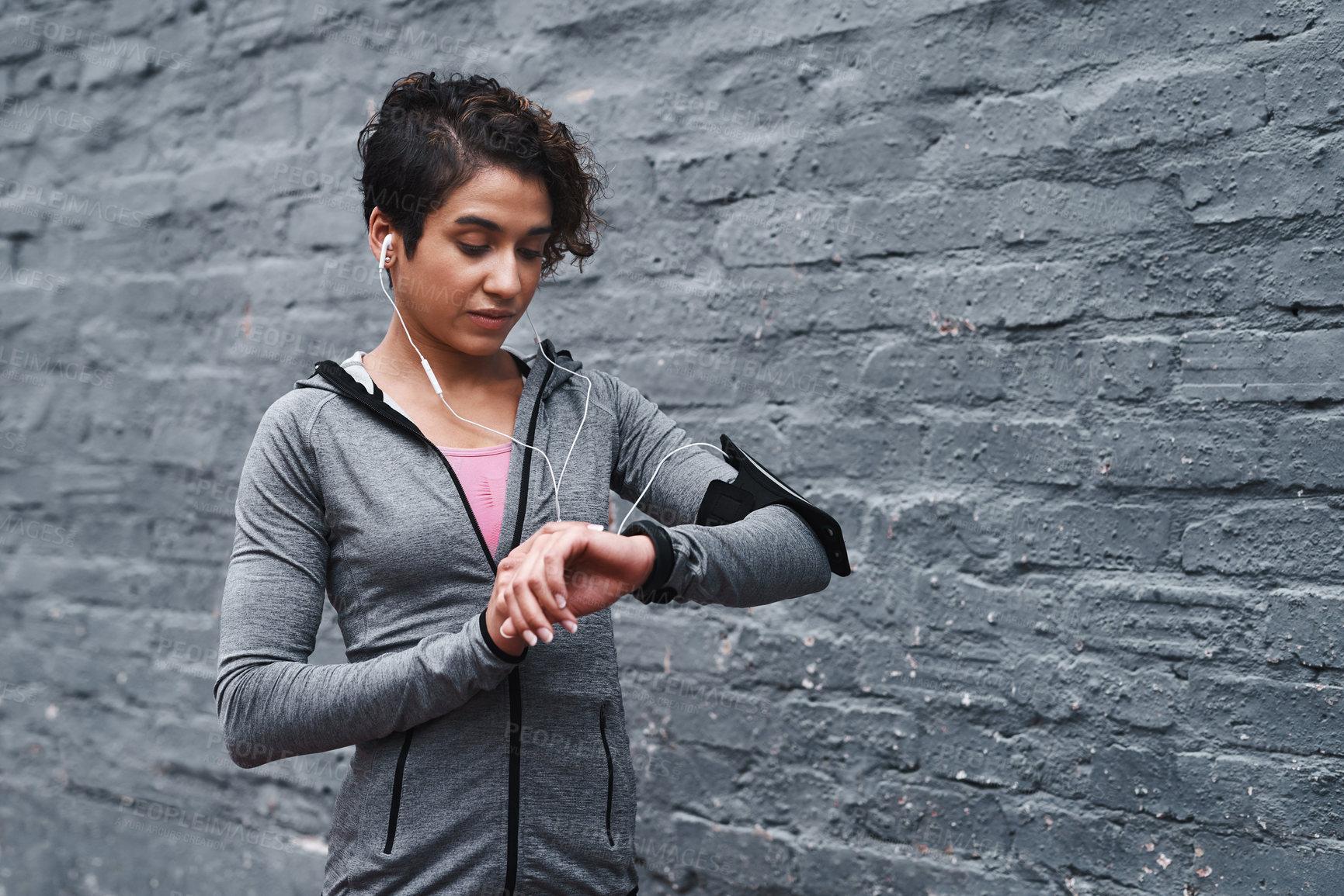Buy stock photo Cropped shot of an attractive young woman wearing earphones and looking at her fitness tracker while standing outside