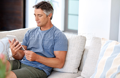 Buy stock photo Cropped shot of a handsome mature man sitting on his living room sofa and using a tablet during the day