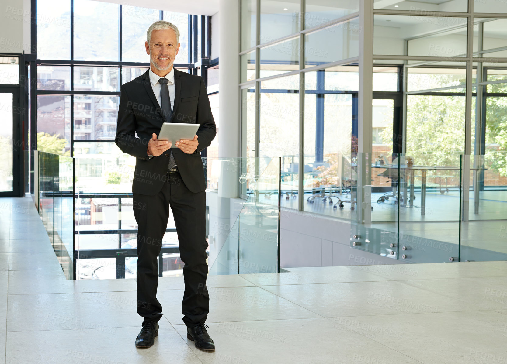 Buy stock photo Full length portrait of a handsome mature businessman standing alone in a modern office and holding a tablet