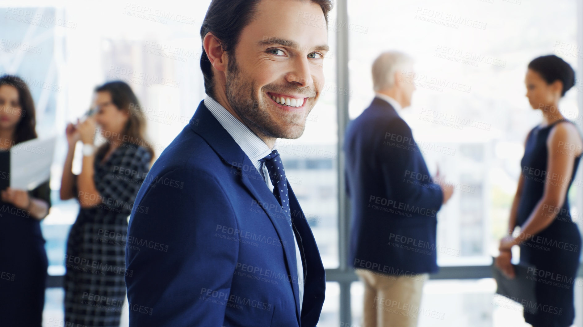 Buy stock photo Shot of a handsome businessman smiling at the camera while colleagues are blurred in the background