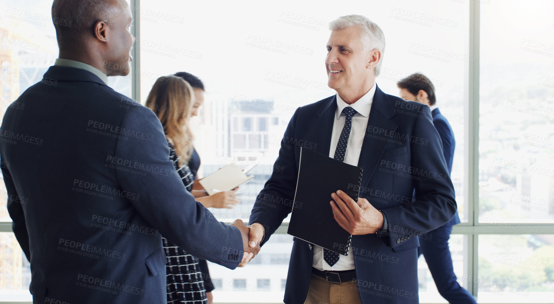 Buy stock photo Cropped shot of two businesspeople shaking hands while colleagues are blurred in the background
