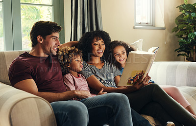 Buy stock photo Shot of a beautiful young family of four reading a story book together while relaxing on a sofa at home